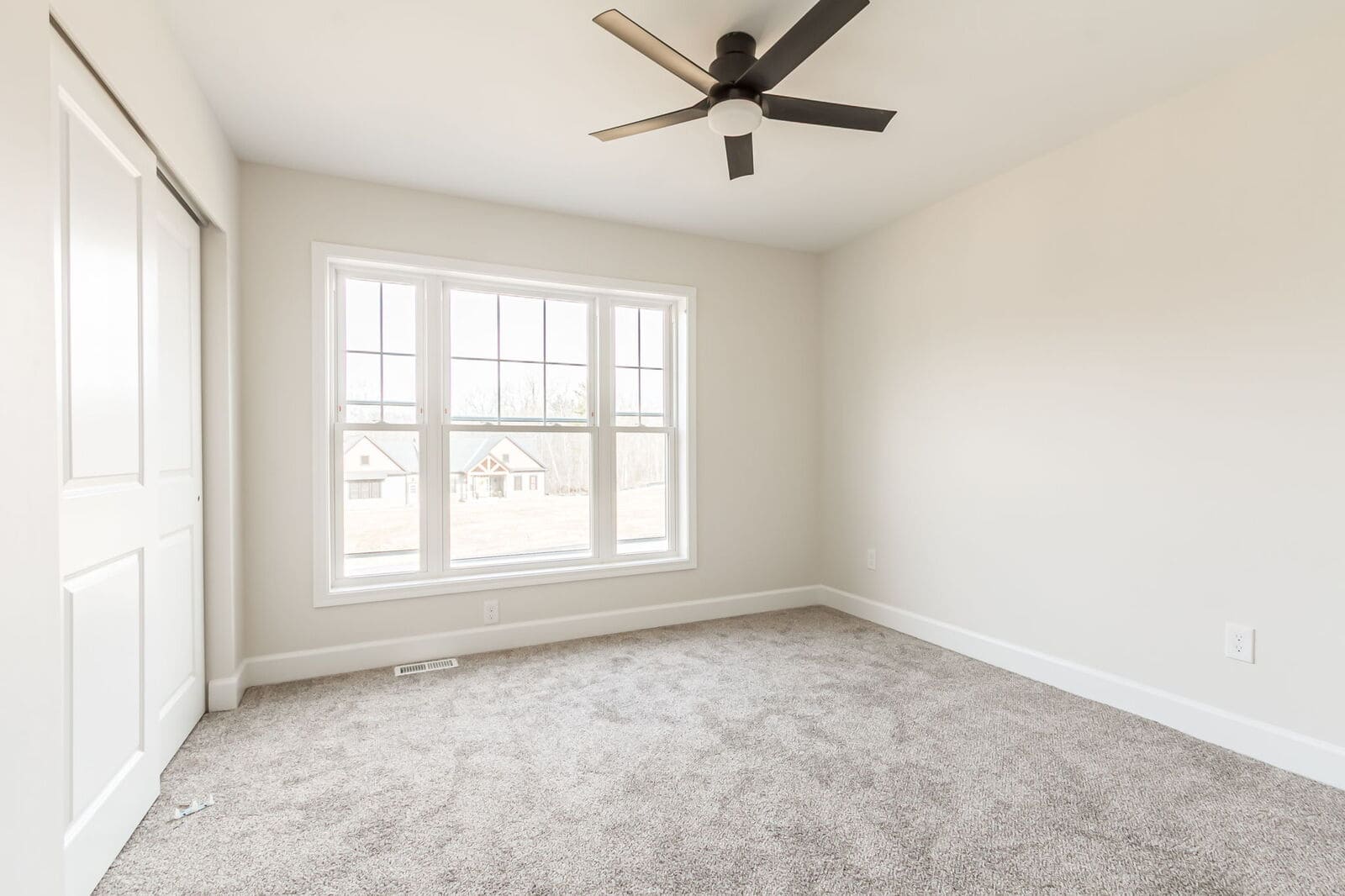 Bright bedroom with neutral walls, gray carpeting, a large window, and a modern black ceiling fan.