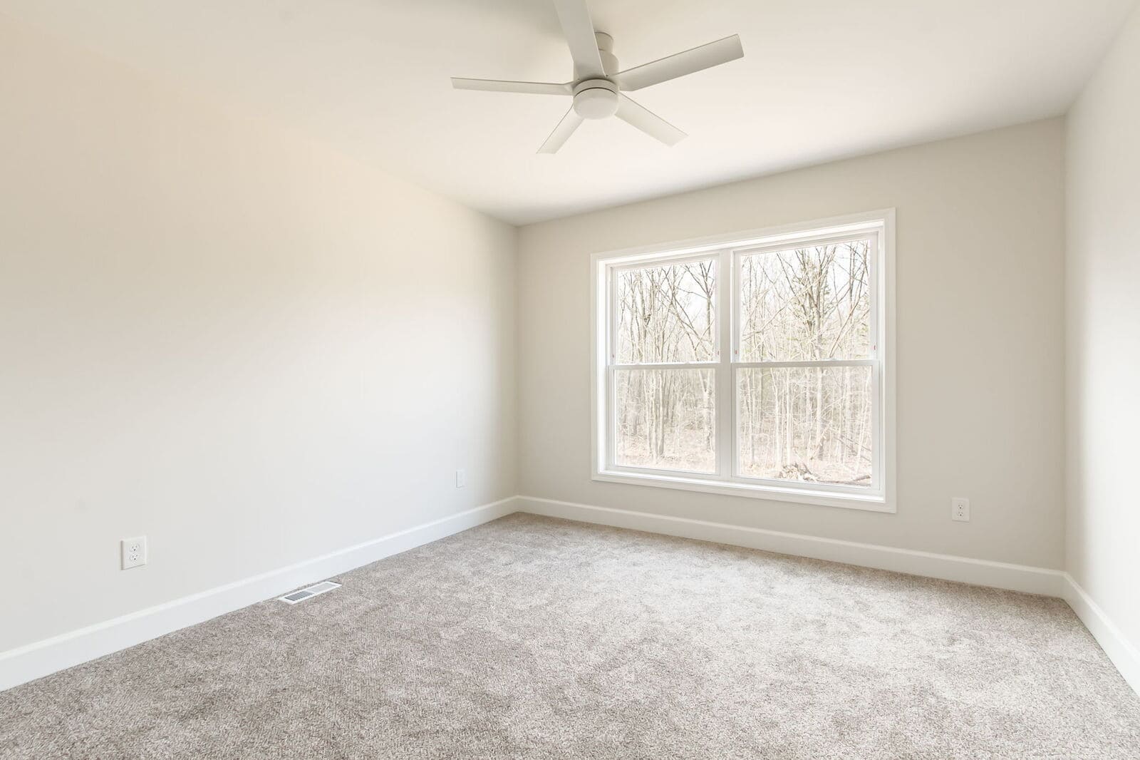 Bright bedroom with soft gray carpeting, a large window overlooking trees, and a white ceiling fan.