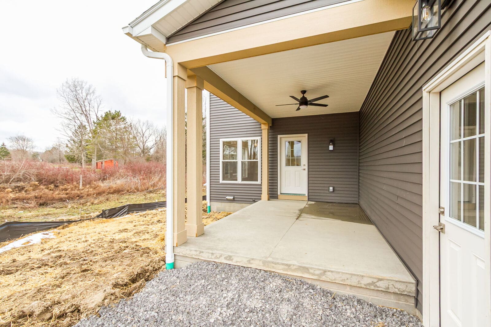 Covered concrete patio with gray siding, tan support posts, exterior doors, and a black ceiling fan.