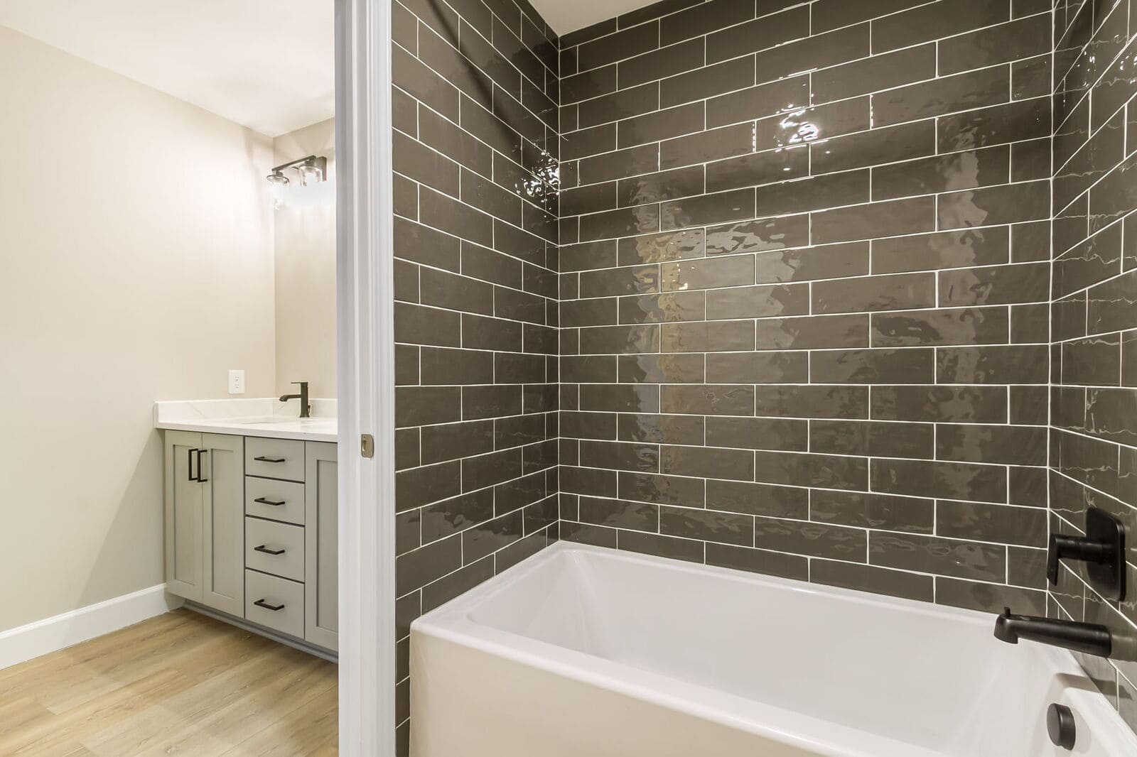 White bathtub with glossy dark subway tile surround, black fixtures, and a gray vanity nearby.