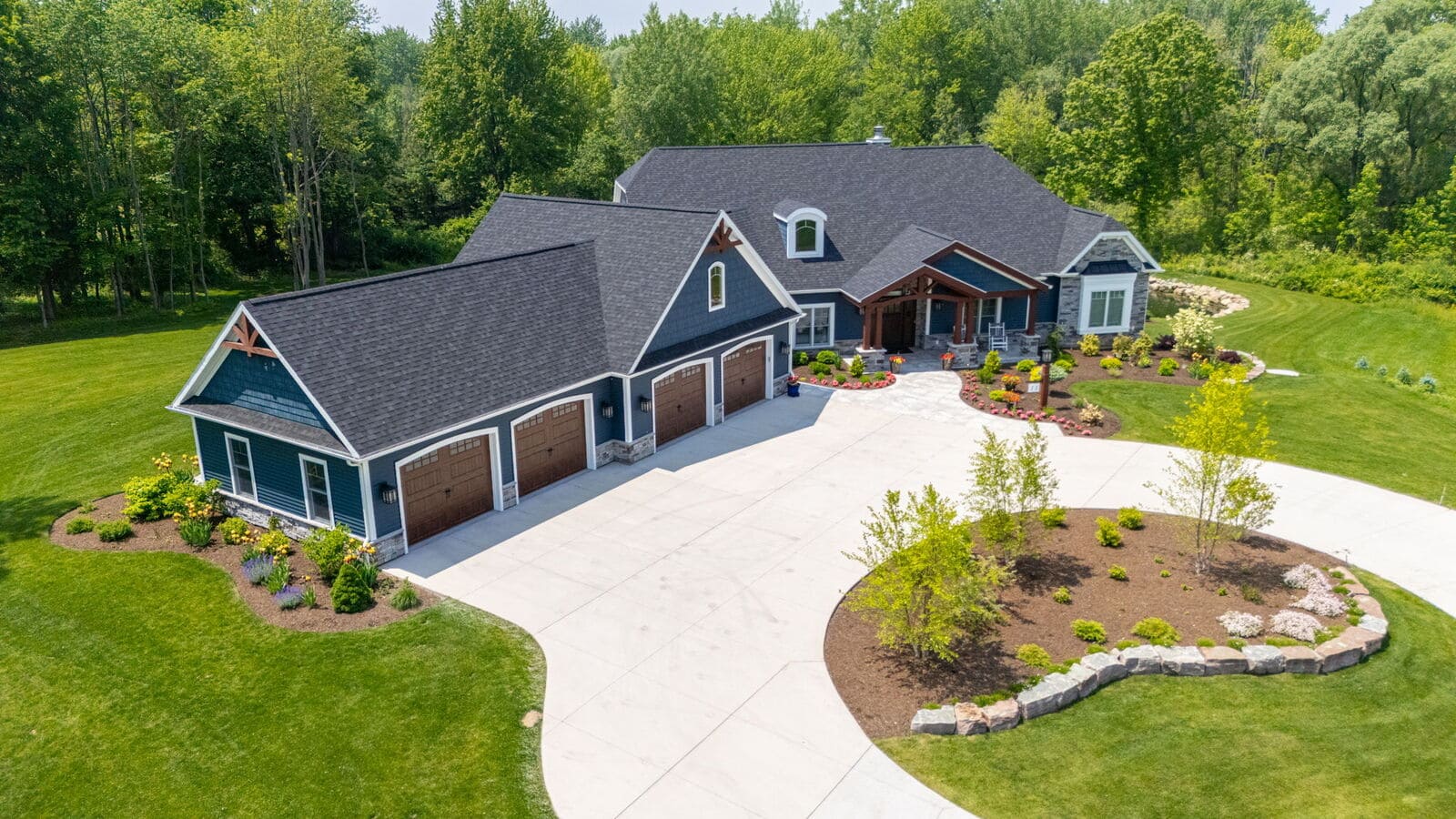 Aerial view of blue custom home with circular driveway, landscaped front yard, and pond in the distance.