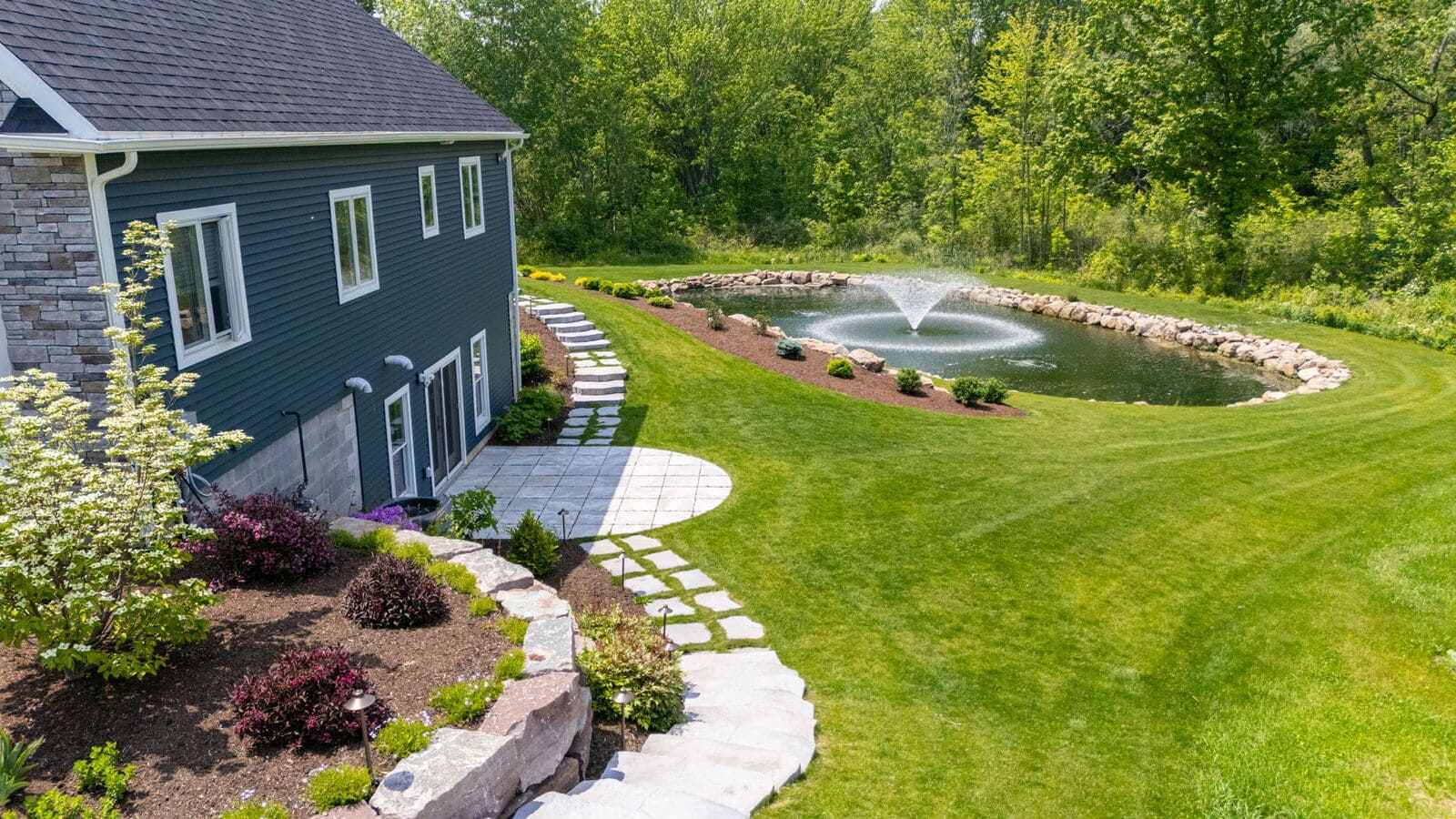 Backyard view with stone walkway, patio, pond fountain, and manicured lawn at a Penfield custom home.