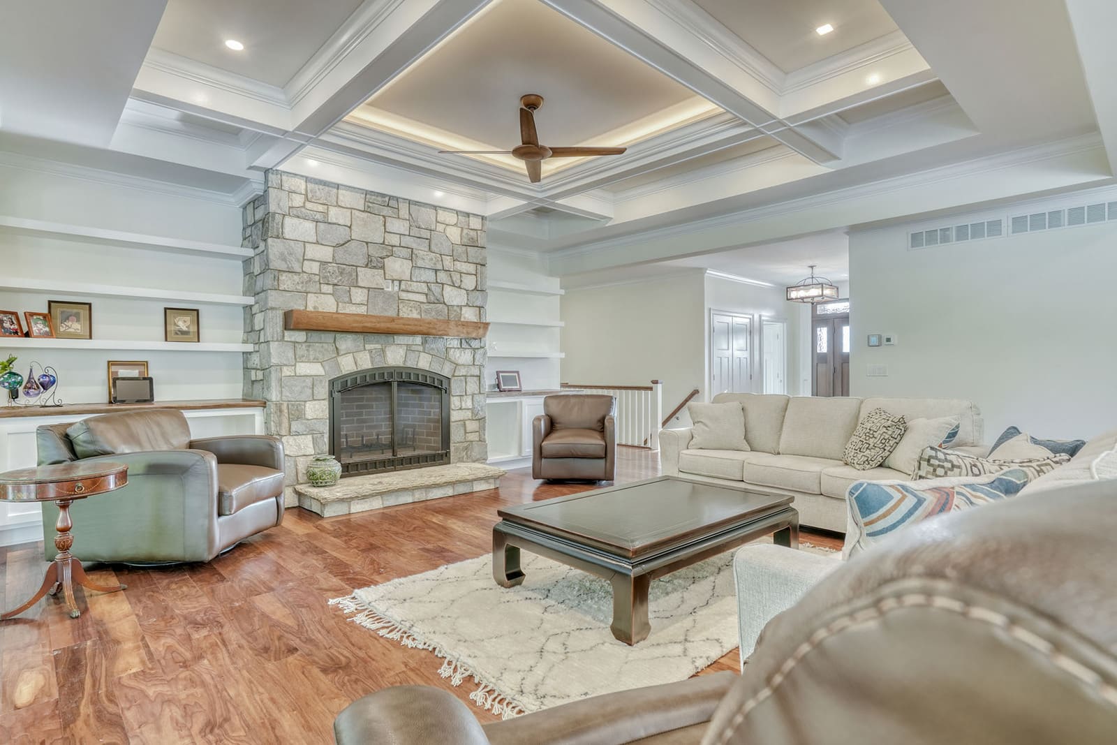Bright living room with stone fireplace, coffered ceiling, built-in shelves, and hardwood floors.