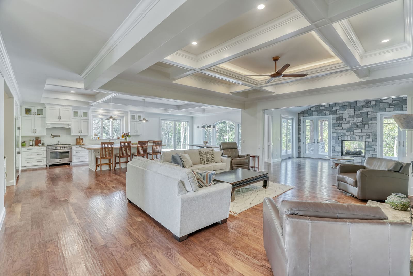 Bright open-concept living area with kitchen, coffered ceilings, stone fireplace, and hardwood floors.