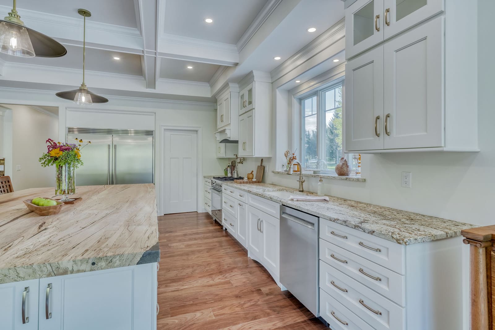 Bright white custom kitchen with coffered ceiling, large island, granite countertops, and wooded views.