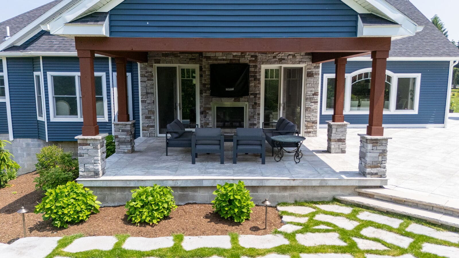 Covered backyard patio with stone columns, outdoor seating, fireplace, and landscaped stone walkway.