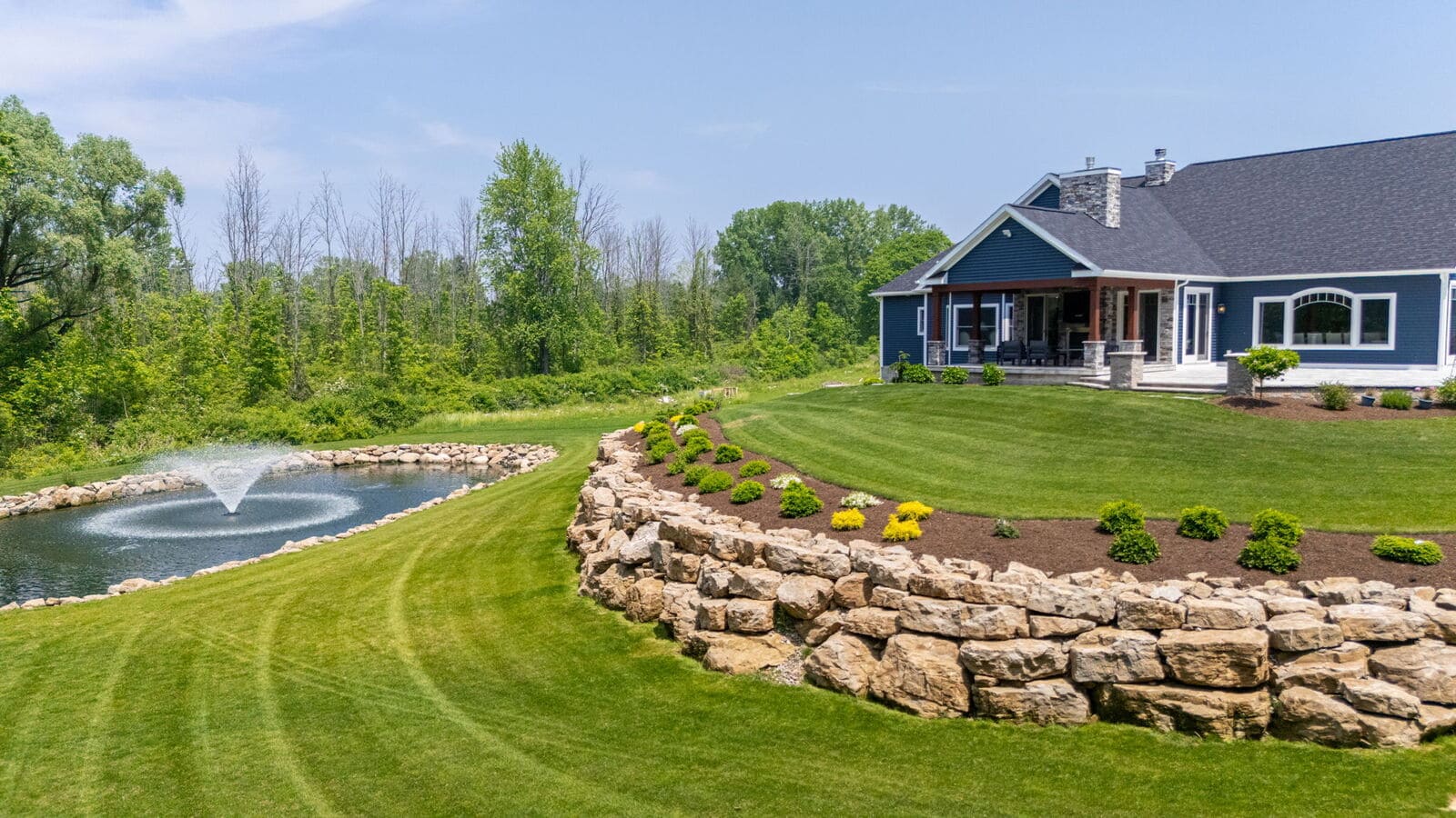 Custom home backyard with pond fountain, stone retaining wall, covered patio, and landscaped lawn.