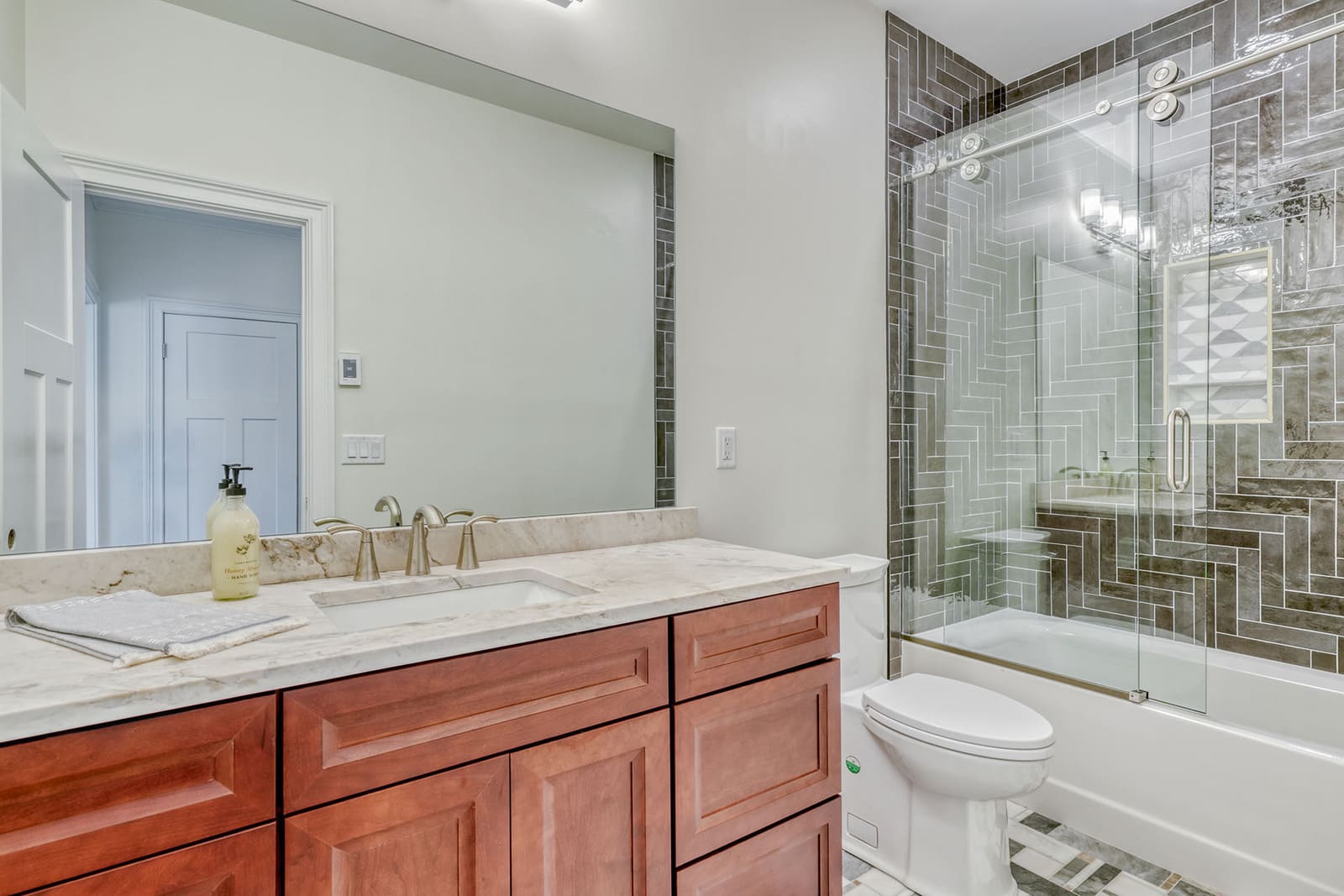Full bathroom with wood vanity, marble counter, large mirror, and glass shower with gray tile.