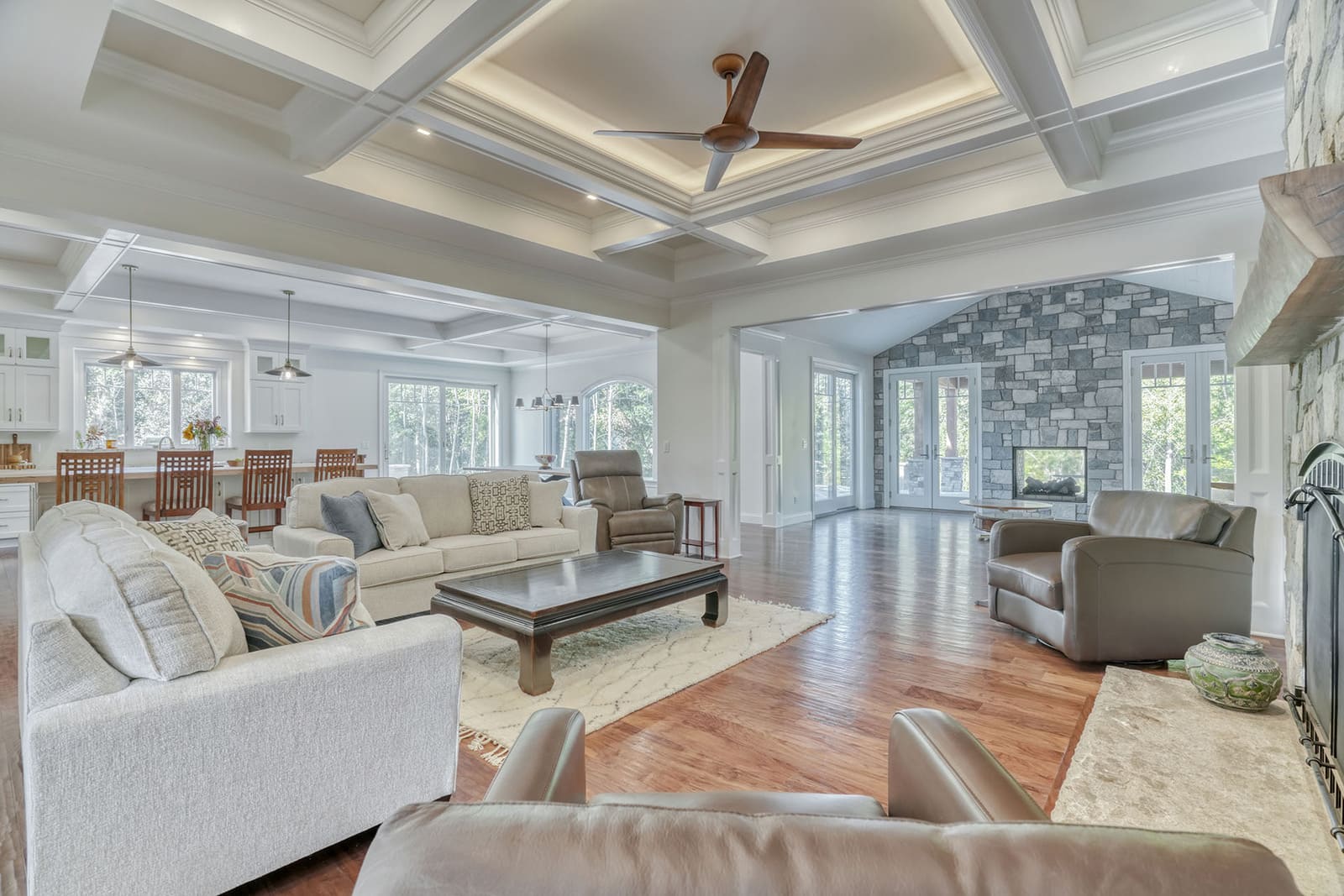 Spacious custom living room with coffered ceiling, open kitchen, stone fireplace, and large windows.