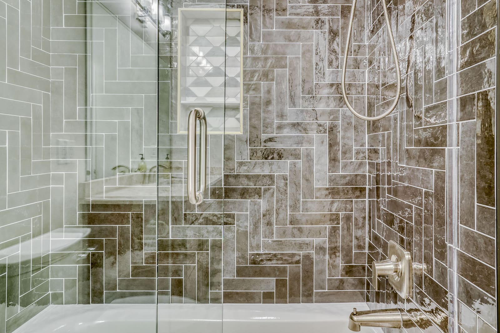 Tile shower with glass enclosure, herringbone wall pattern, built-in niche, and brushed-nickel fixtures.