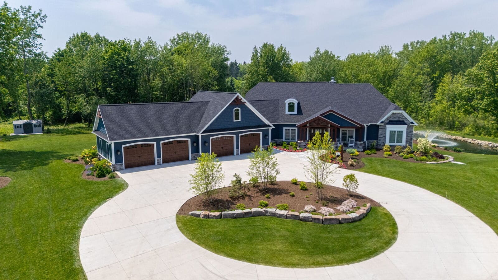 Aerial view of blue custom home with circular driveway, landscaped island, pond, and wooded setting
