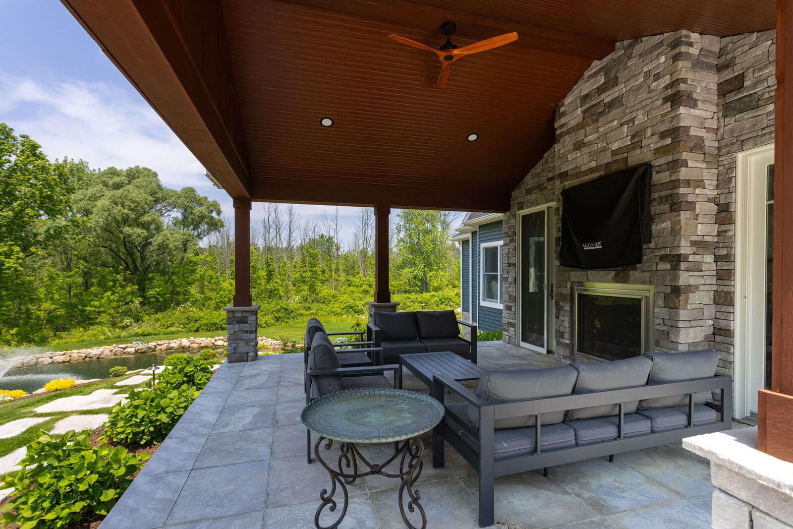 Covered outdoor living space with stone fireplace, wood ceiling, lounge seating, and pond views in Penfield