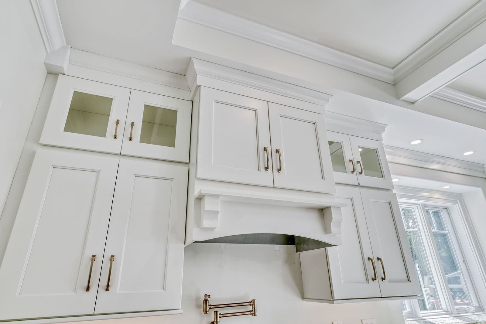 Custom range hood with white cabinetry, glass-front uppers, brass hardware, and coffered ceiling detail