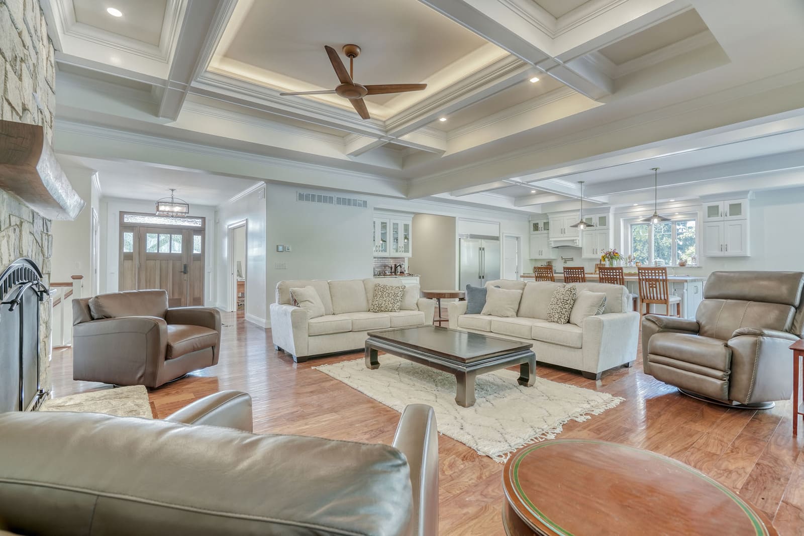 Open living room and kitchen with coffered ceilings, hardwood floors, stone fireplace, and island seating