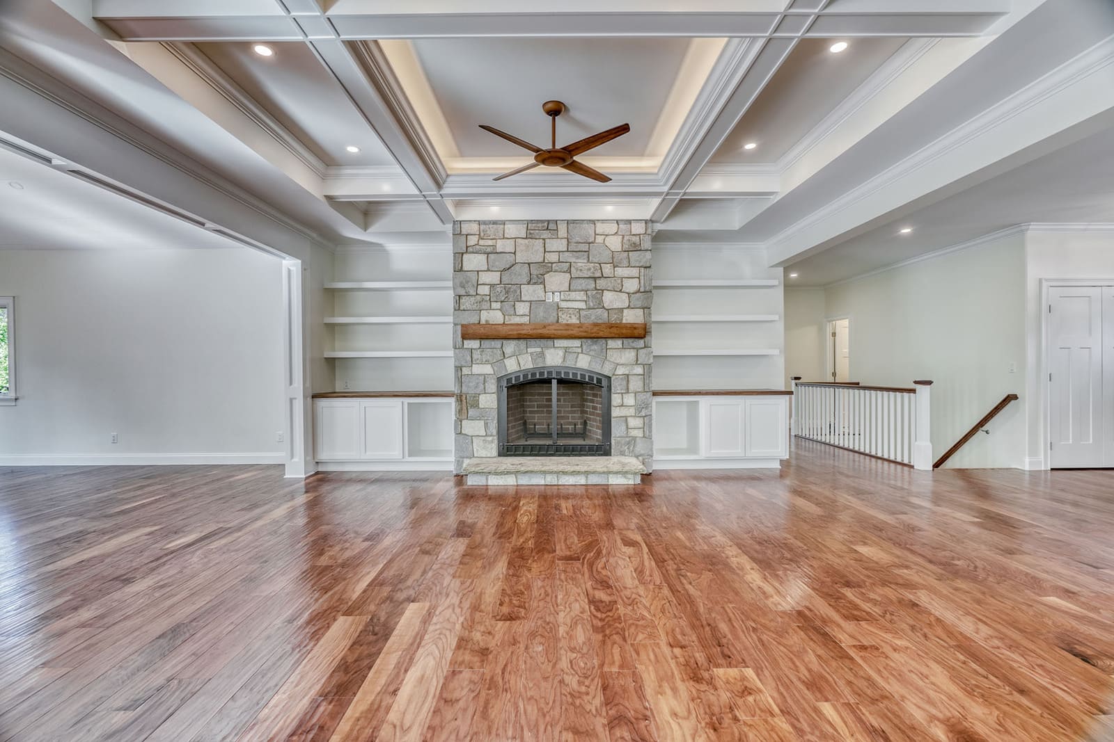 Open living room with stone fireplace, built-in shelves, coffered ceiling, and hardwood floors